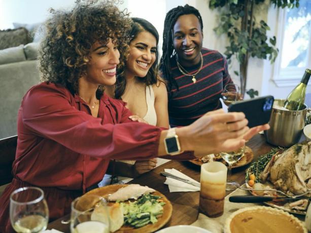 drie vrouwen die een selfie maken boven een eettafel met een emmer met wijn, kalkoen, een pompoentaart, een kaars, borden met eten erop en een wijnglas op tafel