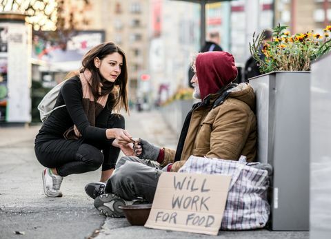 Jonge vrouw die geld geeft aan dakloze bedelaarman zitting in stad.