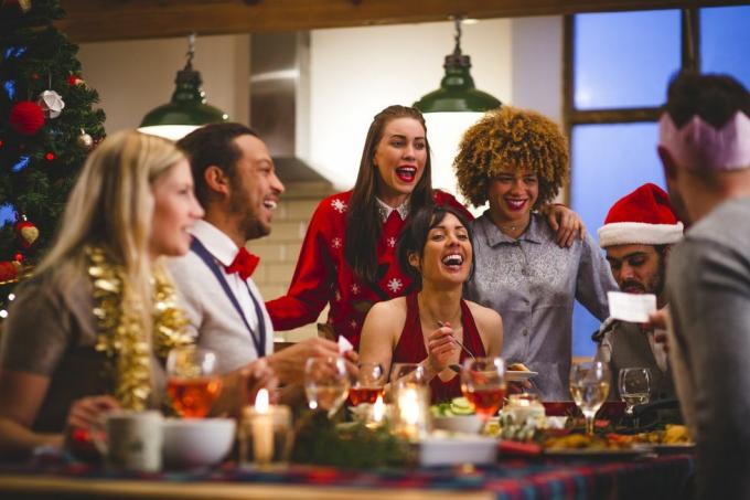 groep vrienden lachen terwijl ze rond een tafel zitten eten en drinken met Kerstmis een man leest een grap voor van een kerstkraker feestmutsen en kersttruien worden gedragen