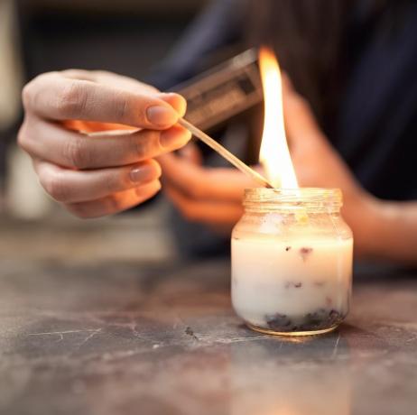anonieme vrouw bijsnijden met brandende lucifer die aromatische kaars in glazen pot aansteekt, thuis op marmeren tafel geplaatst