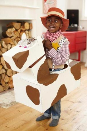 kleine jongen verkleed als cowboy met een cowboyhoed en een geruit hemd en bandana met een kartonnen paard om zijn middel