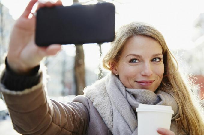 knappe vrouw die in de winter door de straten van de stad loopt, een selfie neemt en een kopje koffie vasthoudt, met achtergrondverlichting