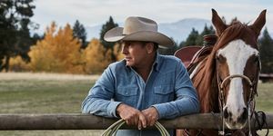 Kevin Costner in Yellowstone naast een paard leunend op een hek met touw in handen, gekleed in een vervaagd blauw spijkerhemd en een beige cowboyhoed