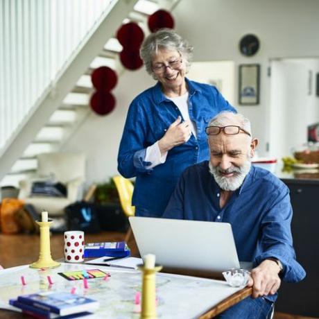 senior vrouw glimlacht en kijkt over de schouder van haar man terwijl hij online recensies onderzoekt en leest over plaatsen om te verblijven en locaties om te bezoeken, plaknotities en gidsen op tafel, vakantie boeken online