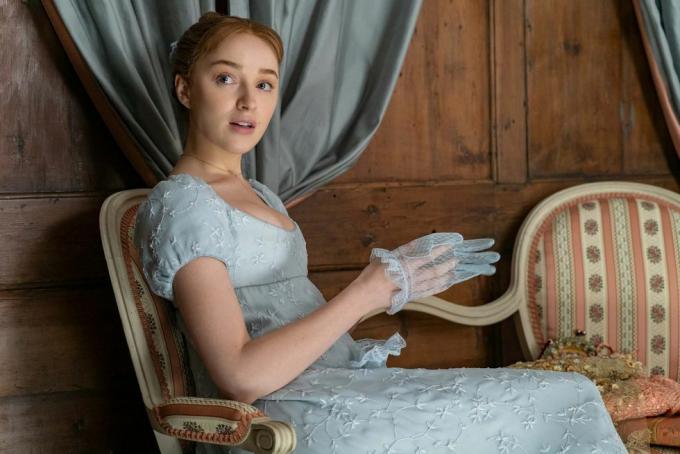 Phoebe Dynevor als Daphne Bridgerton in aflevering 102, zittend, gekleed in een blauwe, rijk getailleerde jurk