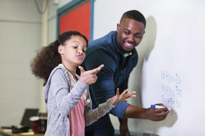 een multiraciaal 9-jarig meisje in wiskundeles, staande bij het whiteboard met haar leraar, een jonge Afro-Amerikaanse man in zijn Jaren '20 kijkt de leraar naar zijn basisschoolleerling, wachtend terwijl ze probeert het antwoord te vinden waarop de nadruk ligt meisje