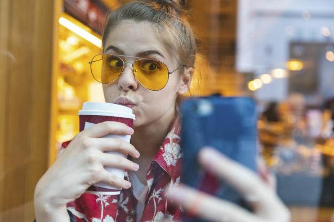 jonge vrouw buiten in zonnebril die een selfie neemt terwijl ze een to go-koffie drinkt