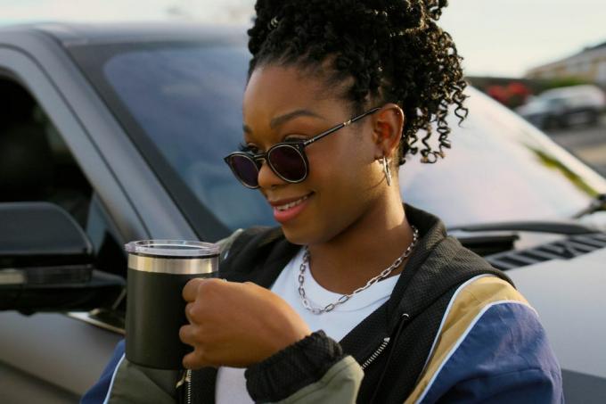 izzy's personage staat naast een lincoln suv een kopje koffie te drinken, met een zonnebril op