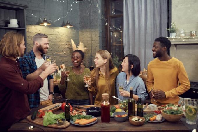 multi-etnische groep glimlachende jonge mensen die samen genieten van een diner, aan tafel staan ​​in een modern interieur en wijnglazen vasthouden